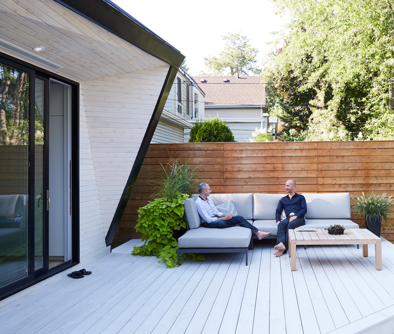 Leslieville backyard patio with white-washed cedar, aluminum, and asymmetrical design, featuring cozy furniture and greenery.